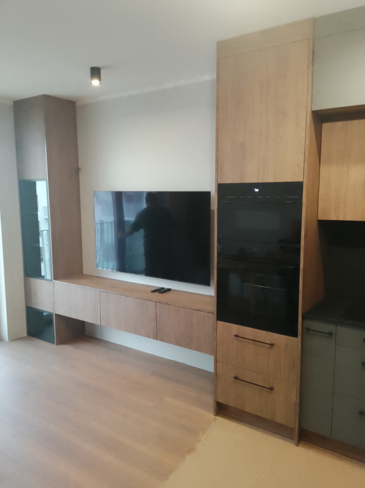 Nowoczesna zabudowa meblowa w salonie z telewizorem i piekarnikiem. Jasne drewno, minimalistyczny design, czarne elementy. W tle widoczne okno.