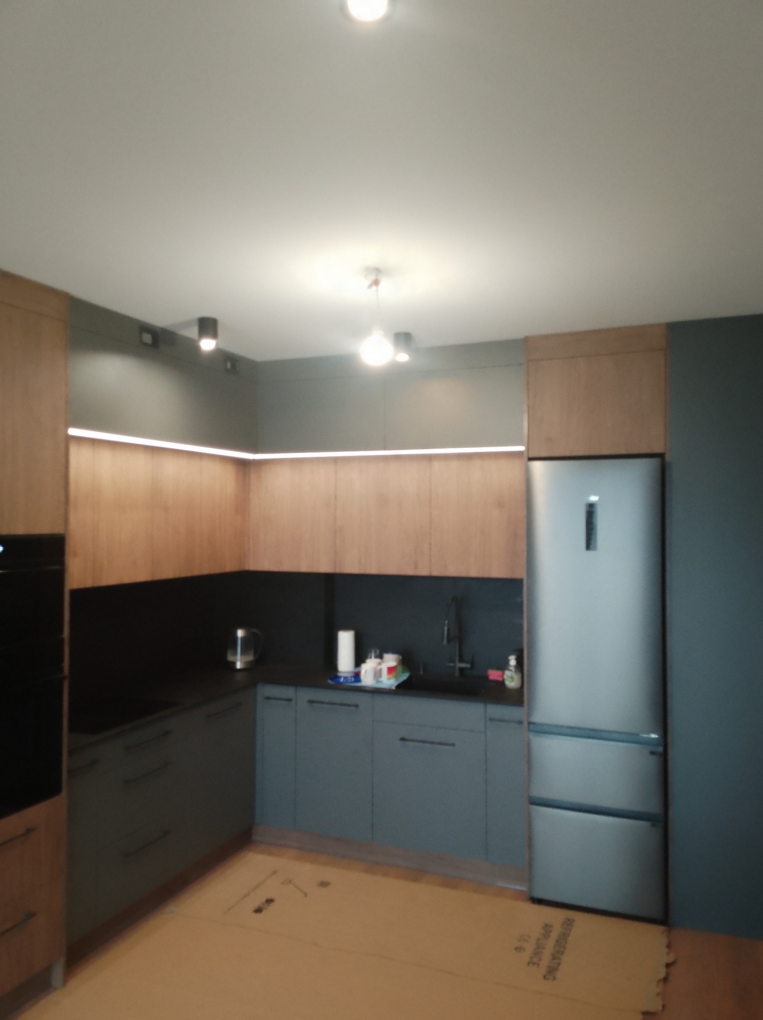 Nowoczesna kuchnia z drewnianymi górnymi szafkami i szarymi dolnymi, oświetlenie LED pod szafkami, srebrna lodówka. Podłoga zabezpieczona kartonem.