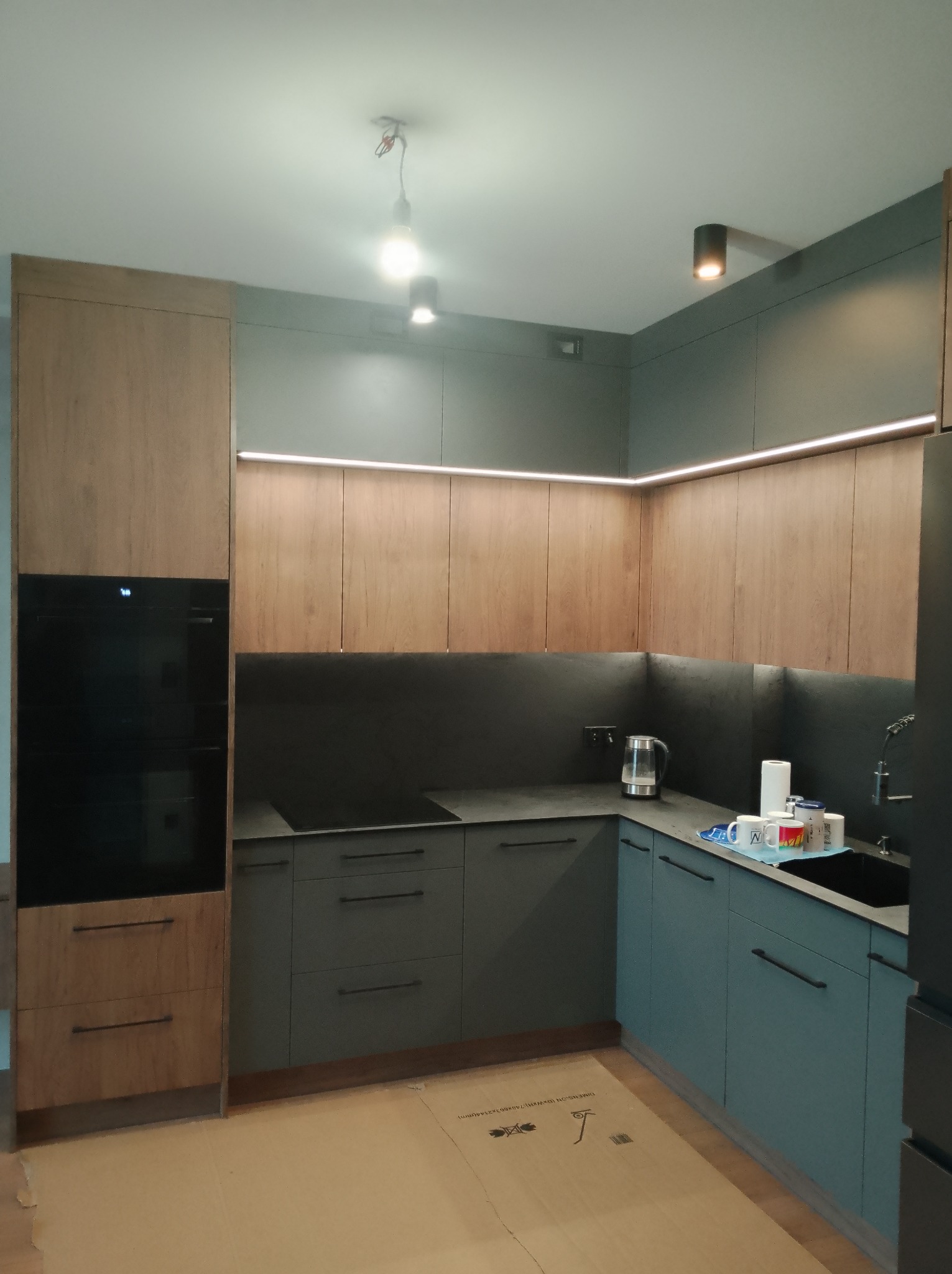 Nowoczesna kuchnia z drewnianymi i szarymi frontami, oświetleniem LED podszafkowym. Uwagę zwraca minimalistyczny design i eleganckie wykończenie. Widoczny fragment blatu i zlewu.