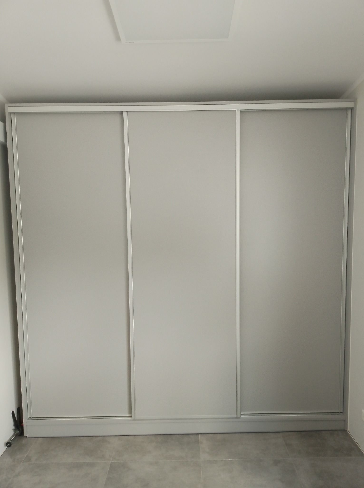 Minimalistyczna szafa wnękowa z trzema przesuwanymi drzwiami w odcieniach szarości, dopasowana do wnętrza z szarą podłogą. Widoczne narzędzia po lewej stronie.