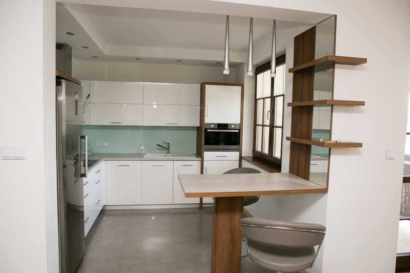 Biała kuchnia z połyskiem, drewniany blat i półki, nowoczesne lampy wiszące. Widok na okno z drewnianą ramą. Minimalistyczny design z elementami drewna.