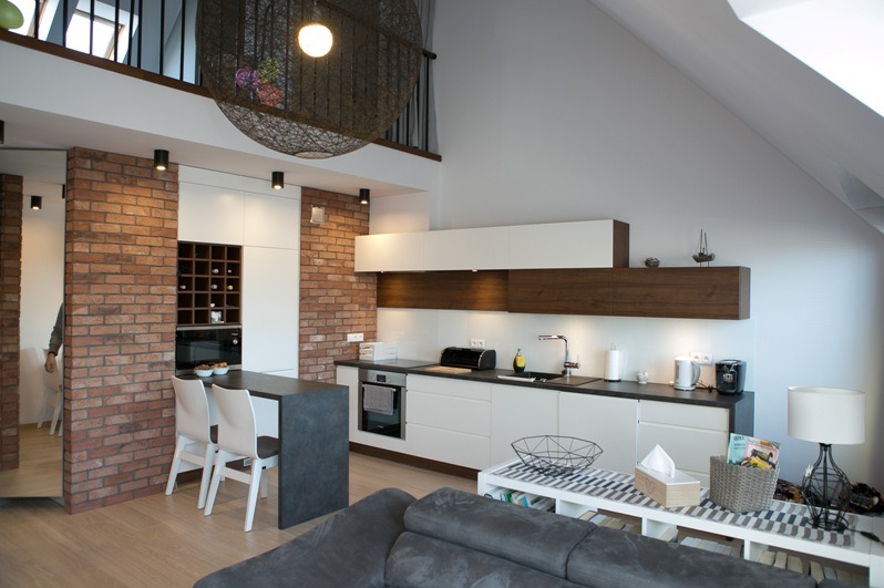Nowoczesna kuchnia w loftowym stylu z ceglaną ścianą, białymi szafkami i drewnianymi akcentami. Nad wyspą wisi dekoracyjna lampa. Widoczny fragment sofy.