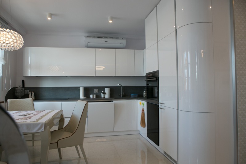 Biała, nowoczesna kuchnia z połyskiem, z zaokrąglonymi szafkami i blatem w odcieniach szarości. Widoczny stół z krzesłami i dekoracyjna lampa.