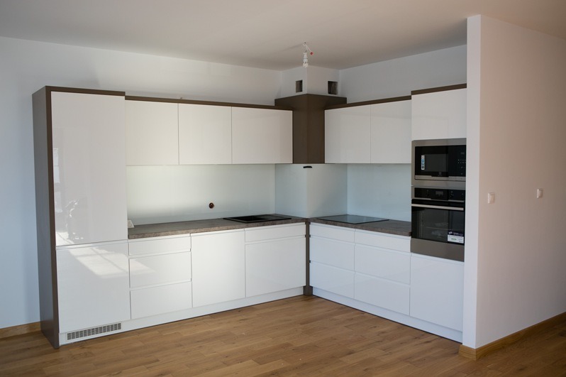 Nowoczesna, narożna kuchnia z białymi, lakierowanymi frontami i ciemnobrązowymi akcentami. Widoczny sprzęt AGD w zabudowie i drewniana podłoga.
