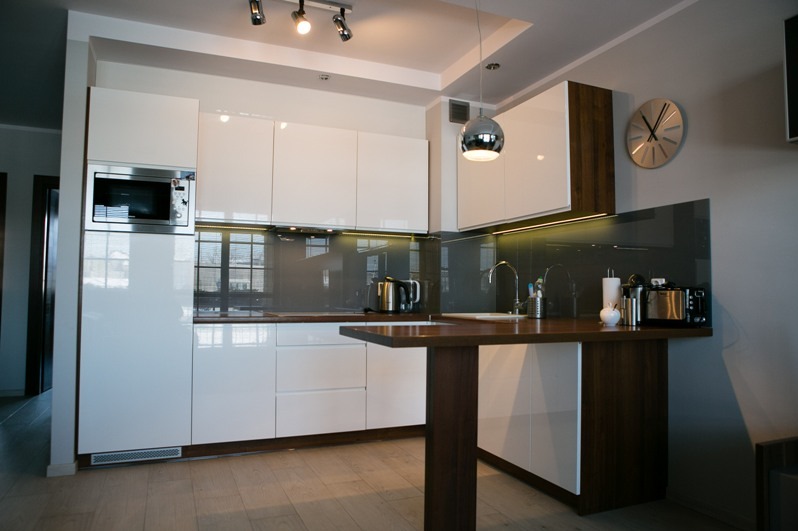 Biała kuchnia na wymiar z drewnianym blatem i barkiem, oświetleniem LED podszafkowym i srebrnym zegarem na ścianie. Nowoczesny design z połyskiem.