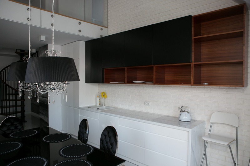 Nowoczesna kuchnia z białymi szafkami dolnymi i czarnymi górnymi, drewniane półki, biała cegła na ścianie, czarny stół i krzesła. Widoczny fragment schodów.