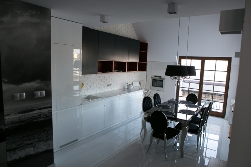 Nowoczesna kuchnia z białymi, błyszczącymi frontami i czarnymi szafkami wiszącymi. Na ścianie fototapeta z motywem morza. Biała podłoga z połyskiem.