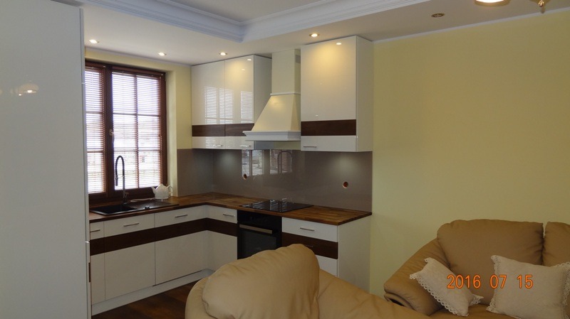 Jasna kuchnia na wymiar z białymi szafkami i drewnianymi akcentami, widok na okno z żaluzjami i fragment sofy w tle. Nowoczesny design z oświetleniem punktowym.