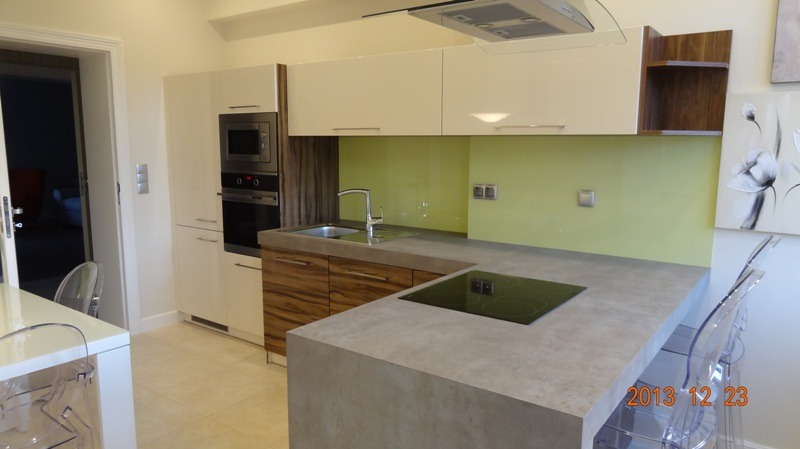 Nowoczesna kuchnia z białymi szafkami, drewnianymi akcentami i blatem imitującym beton. Zielony panel nad blatem i minimalistyczny design.