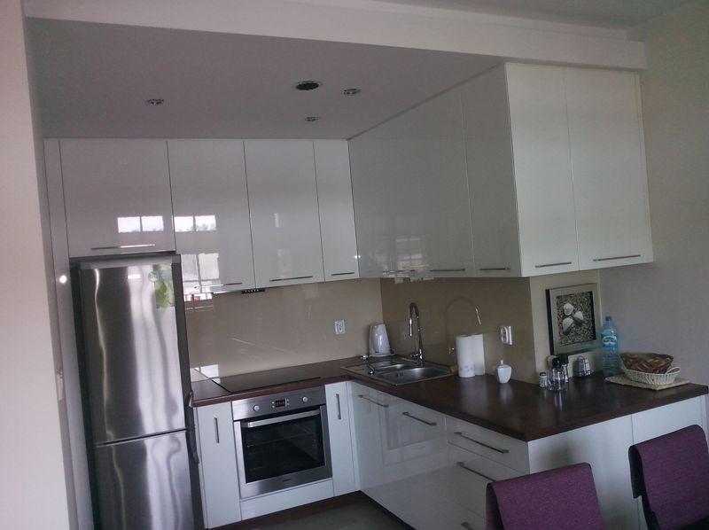 Biała, narożna kuchnia z połyskiem, z zabudową pod sufit i brązowym blatem. Widoczna lodówka ze stali nierdzewnej, piekarnik i zlewozmywak. Ujęcie z perspektywy pomieszczenia.