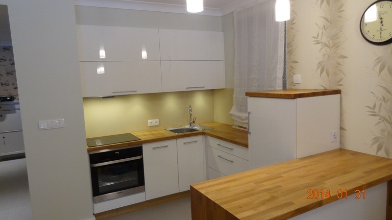 Jasna kuchnia z białymi, lakierowanymi szafkami i drewnianym blatem. Widoczny piekarnik, zlew i wyspa kuchenna. Na ścianie zegar i tapeta w roślinny wzór.