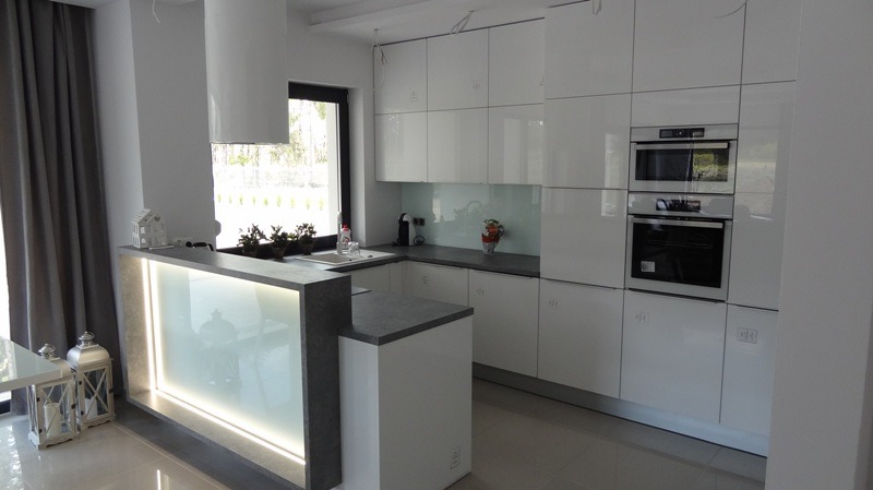 Biała, nowoczesna kuchnia z wyspą i oświetleniem LED. Błyszczące fronty szafek, szary blat, okap cylindryczny. Widok na ogród przez okno.