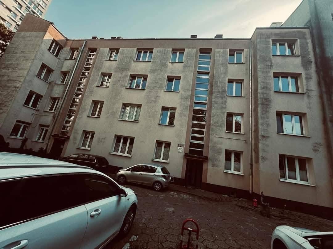 Szary blok mieszkalny z wieloma oknami, widziany z dołu, z zaparkowanymi samochodami. Budynek ma charakterystyczne pionowe wnęki.