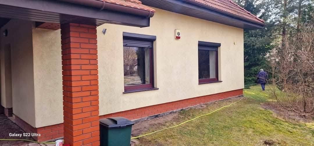 Elewacja domu z dwoma oknami z roletami zewnętrznymi, ceglana podmurówka i filar, trawnik z zieloną trawą. Widoczny system alarmowy na ścianie.