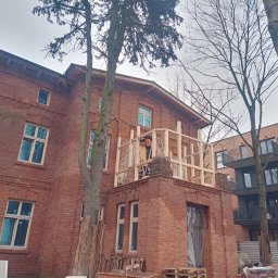 MK Nadzory Budowlane Marek Krajza - Remontowany budynek z czerwonej cegły, z drewnianą konstrukcją na balkonie i pracownikiem na drabinie. Widoczne drzewa i sąsiedni budynek.