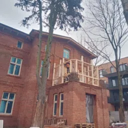 Remontowany budynek z czerwonej cegły, z drewnianą konstrukcją na balkonie i pracownikiem na drabinie. Widoczne drzewa i sąsiedni budynek.