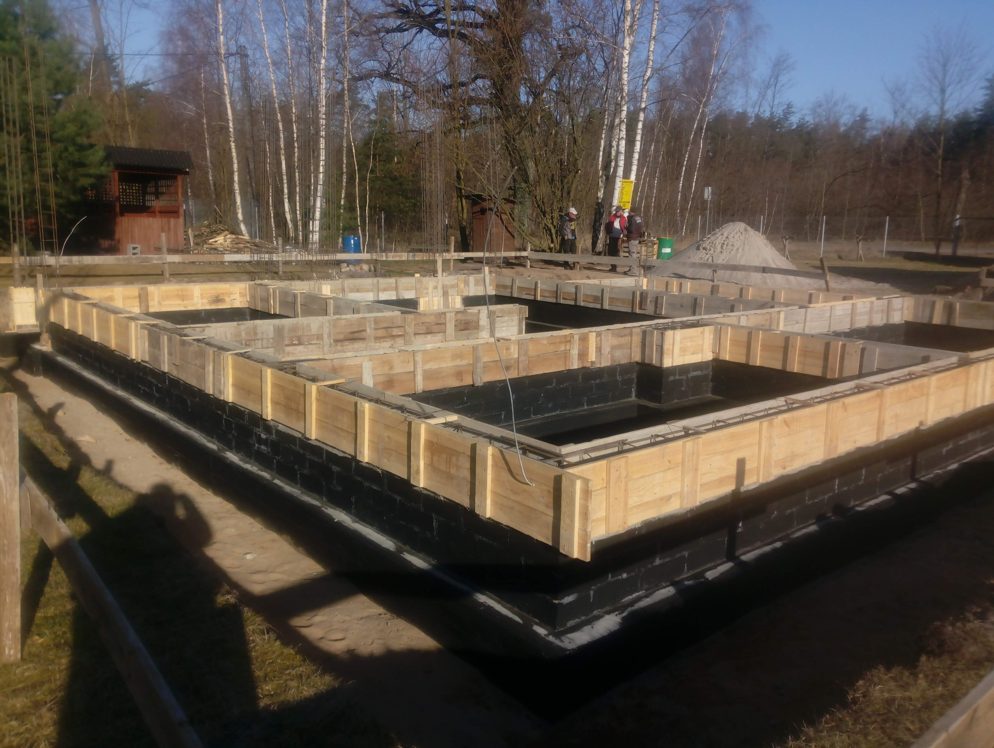 Fundamenty domu jednorodzinnego z drewnianym szalunkiem, izolacją przeciwwilgociową i zbrojeniami. W tle drzewa i ekipa budowlana.