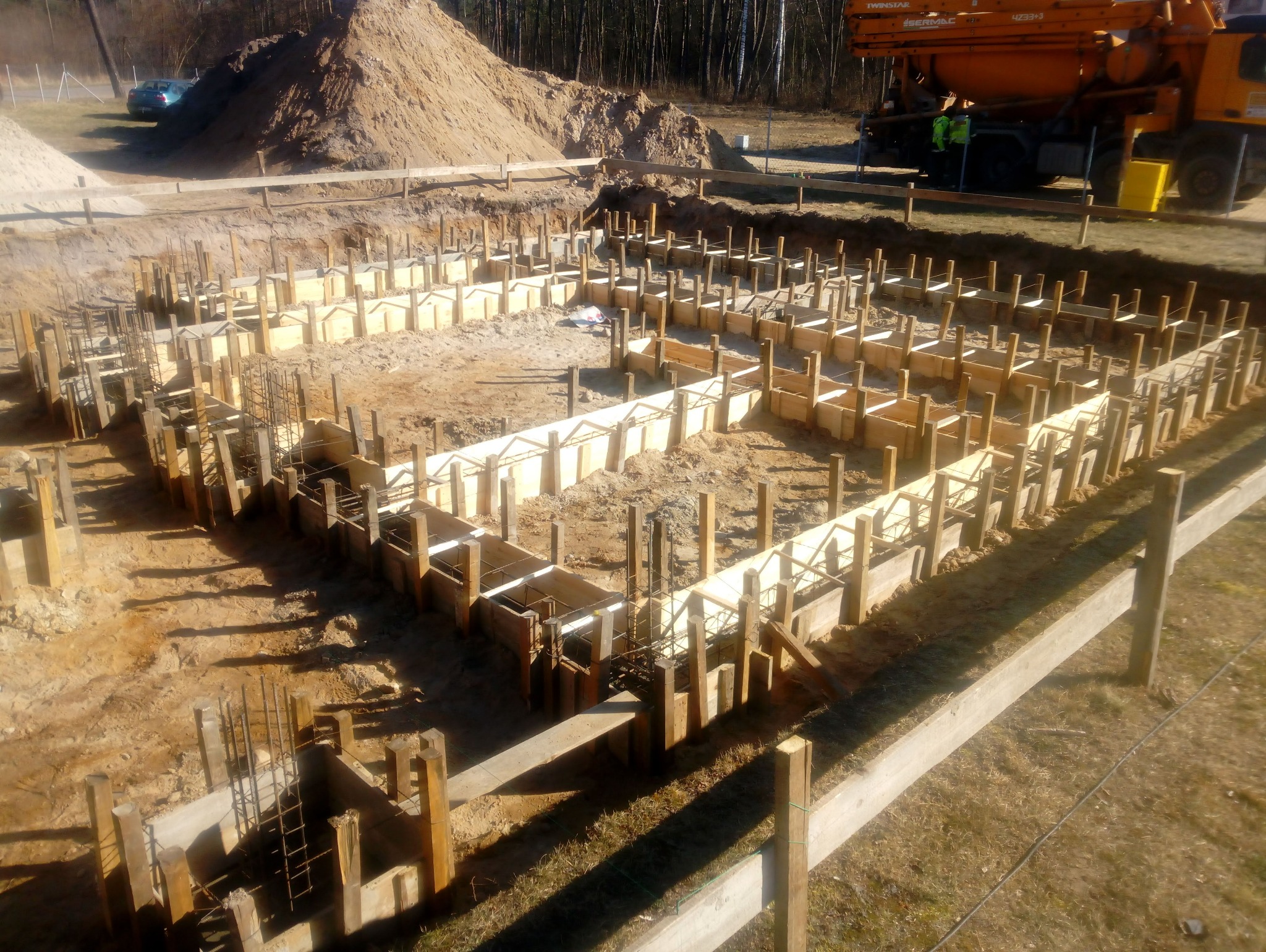 Przygotowanie fundamentów pod dom: drewniany szalunek, zbrojenie stalowe, kopiec piasku w tle, betoniarka. Widok z góry, wczesna faza budowy, słoneczny dzień.