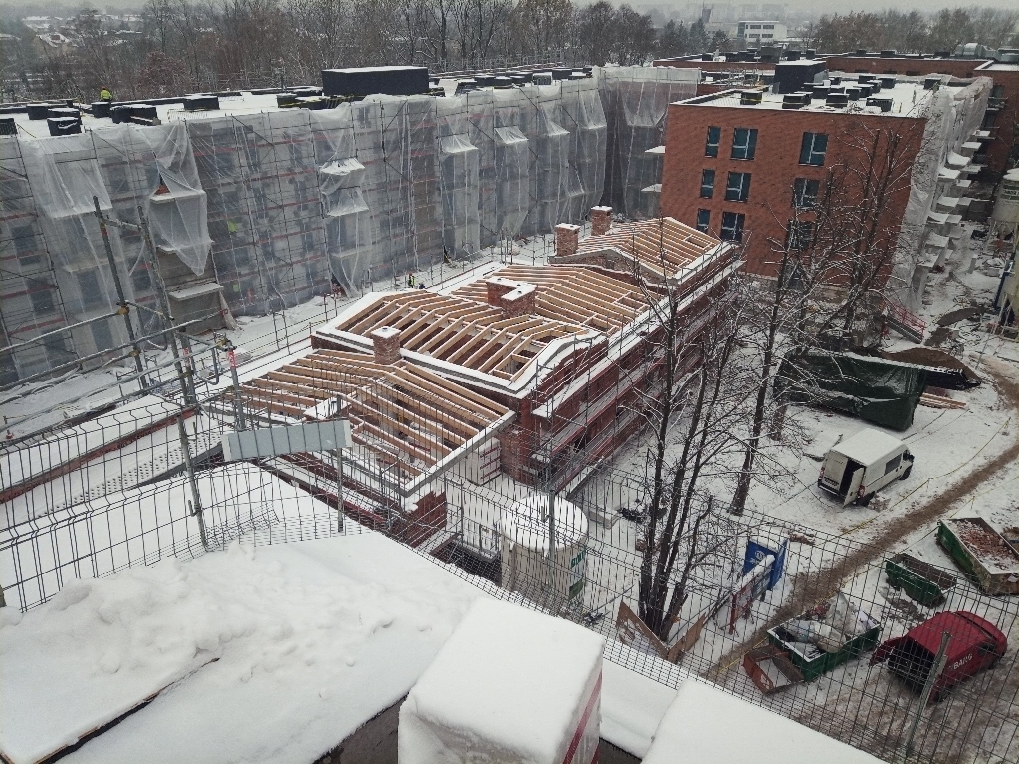 Budowa bloku z czerwonej cegły w zimowej scenerii. Widoczne rusztowania, niezadaszony dach z kominami i zasypane śniegiem otoczenie budowy z kontenerami.