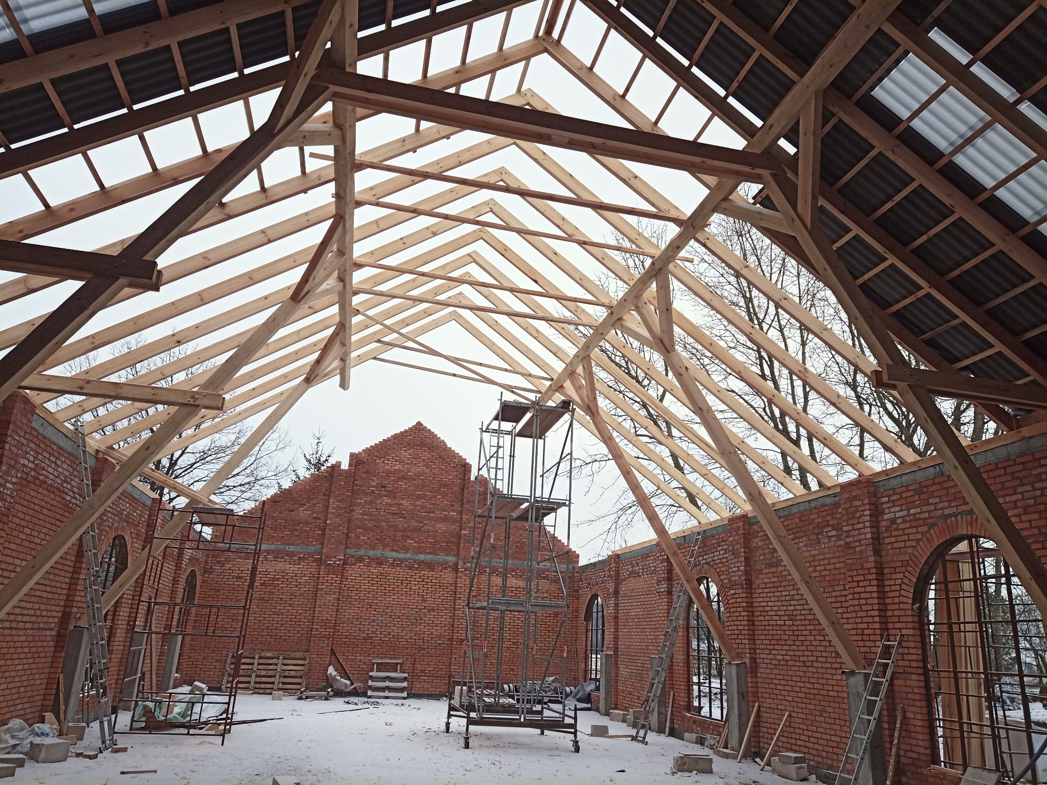 Wnętrze murowanego budynku w trakcie budowy z widoczną konstrukcją drewnianą dachu, rusztowaniami i ceglanymi ścianami z oknami w kształcie łuków.