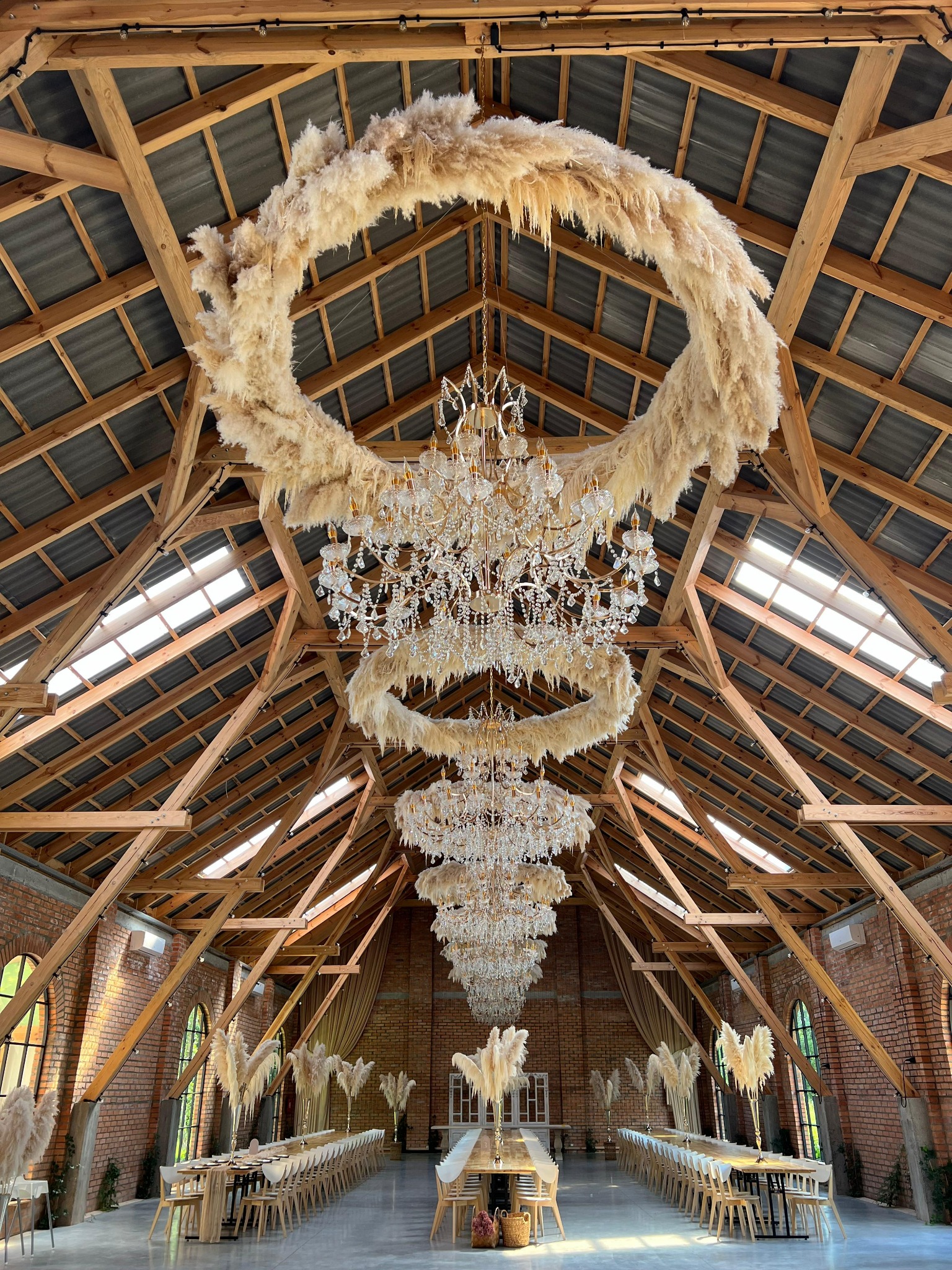 Dekoracja sali weselnej z drewnianą konstrukcją dachu, żyrandolami i okrągłymi dekoracjami z trawy pampasowej. Długie stoły z krzesłami ustawione w rzędach.