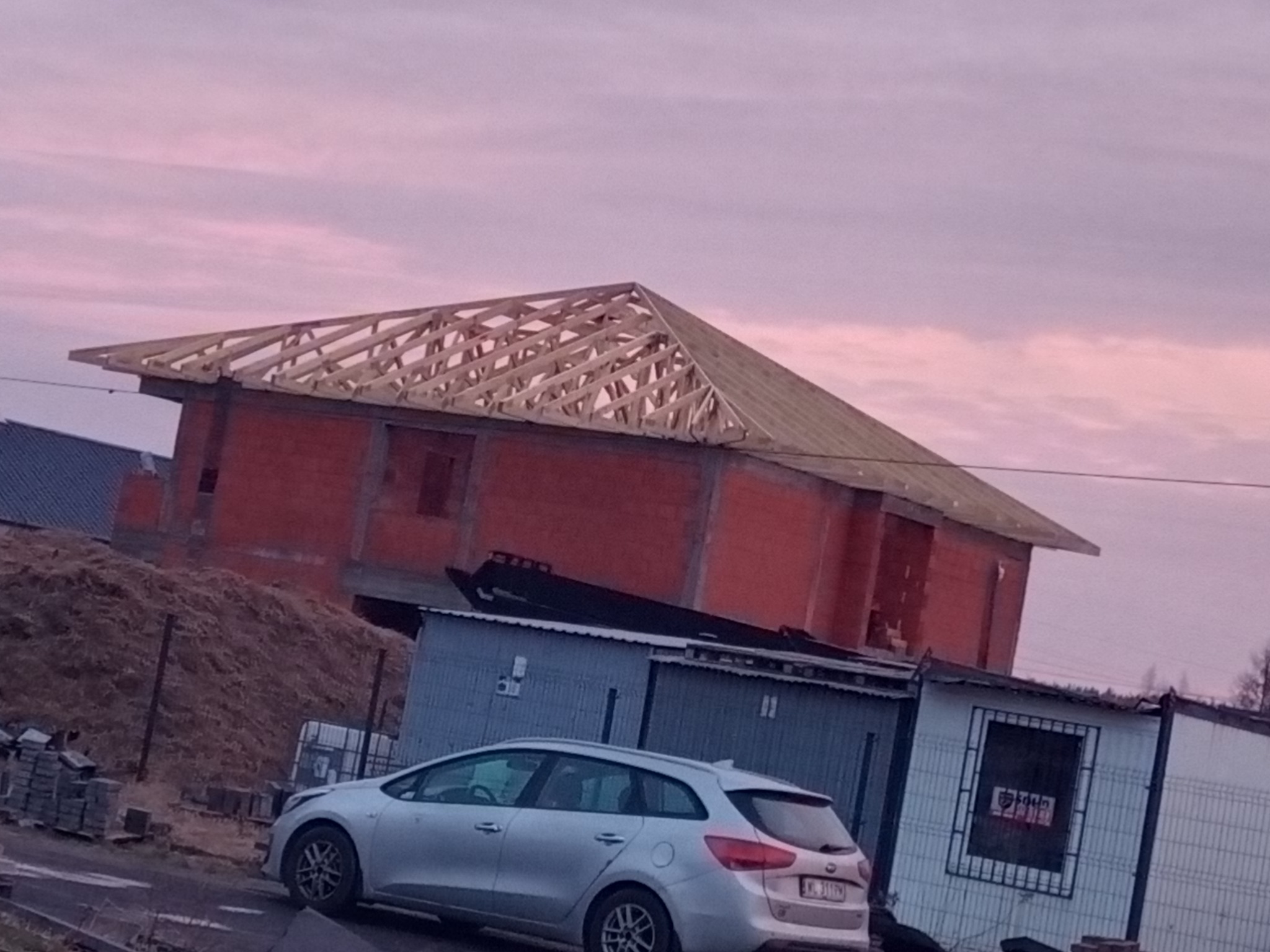 Wznoszony dom z czerwonej cegły z drewnianą konstrukcją dachu w trakcie budowy, widoczny zza zaparkowanego srebrnego kombi i baraków. Niebo w odcieniach różu.