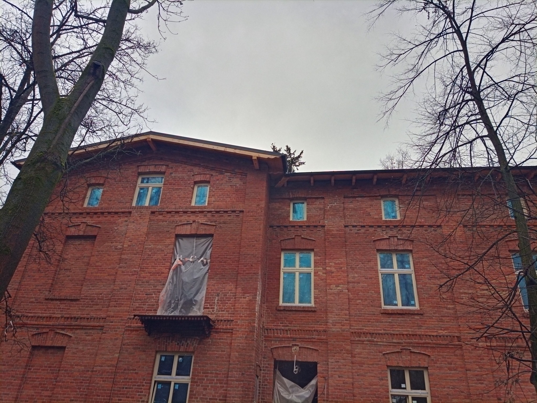 Ceglany budynek w Ostrołęce, zabezpieczony folią podczas prac remontowych. Widoczne okna i elementy architektoniczne. Drzewa bezlistne na pierwszym planie.
