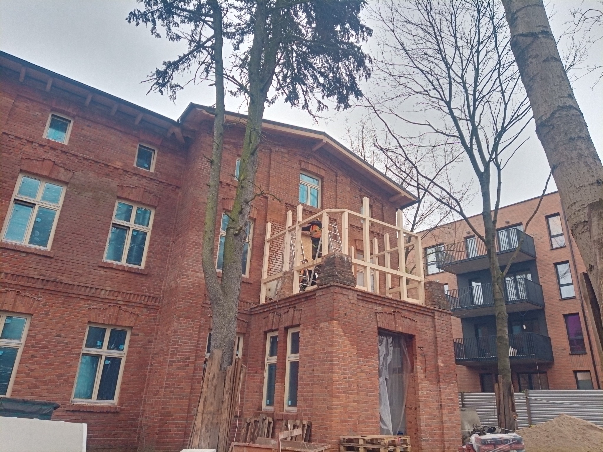 Remontowany budynek z czerwonej cegły, z drewnianą konstrukcją na balkonie i pracownikiem na drabinie. Widoczne drzewa i sąsiedni budynek.