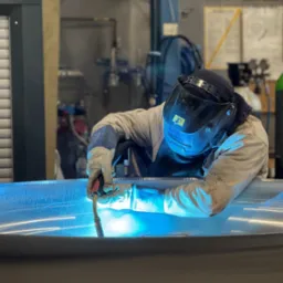 Spawacz w masce spawalniczej łączy metalowy element metodą MIG/MAG. W tle butla z gazem i narzędzia. Widoczne iskry i intensywne światło łuku spawalniczego.