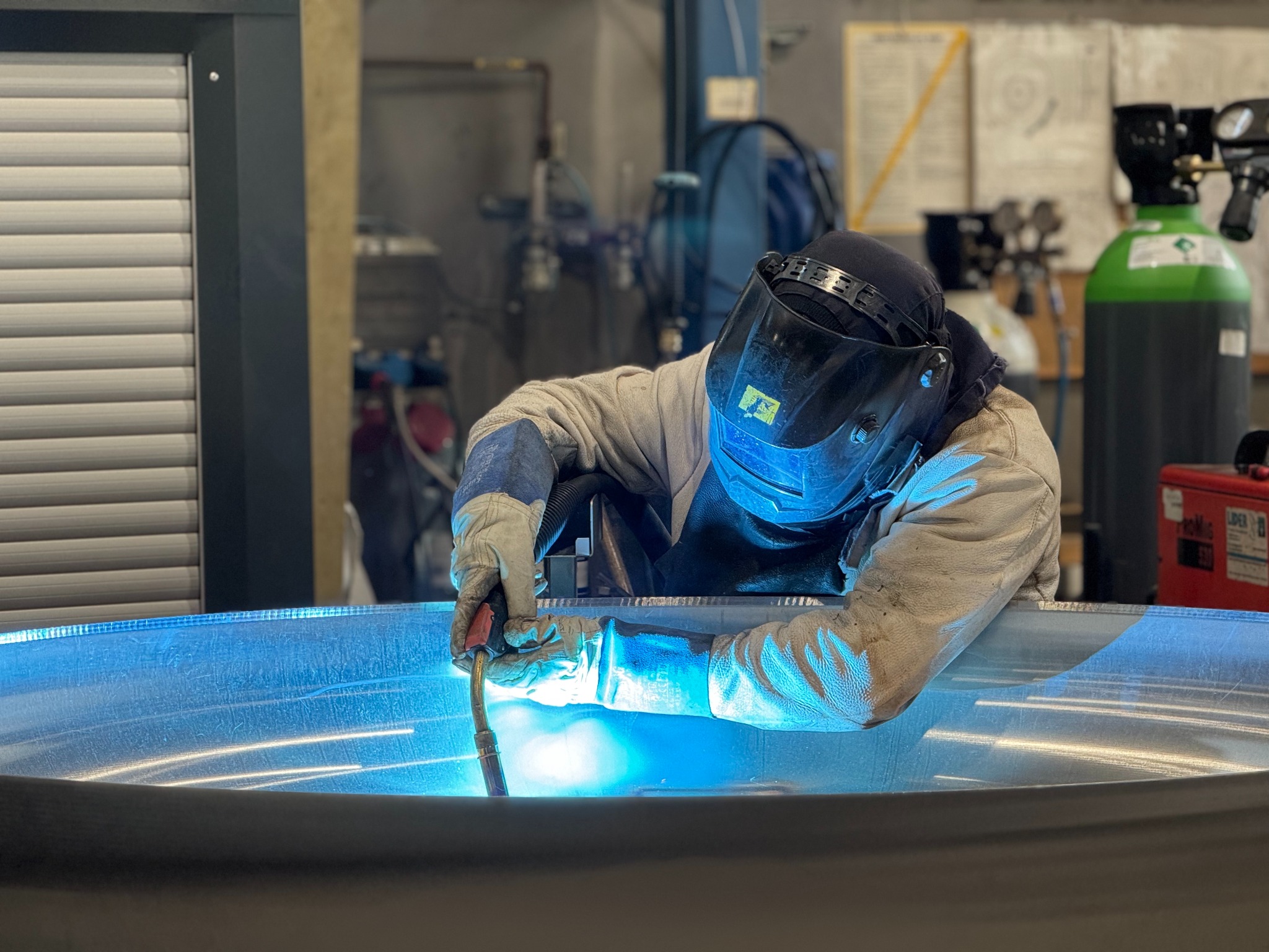 Spawacz w masce spawalniczej łączy metalowy element metodą MIG/MAG. W tle butla z gazem i narzędzia. Widoczne iskry i intensywne światło łuku spawalniczego.