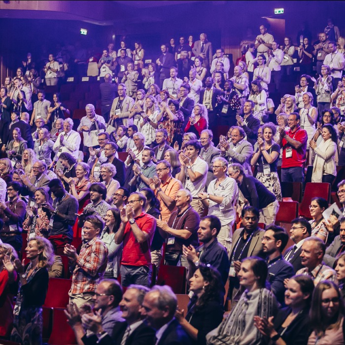 Widok z dołu na widownię podczas wydarzenia. Publiczność klaszcze, oświetlenie sceniczne tworzy atmosferę. Różnorodne stroje i wyrazy twarzy.