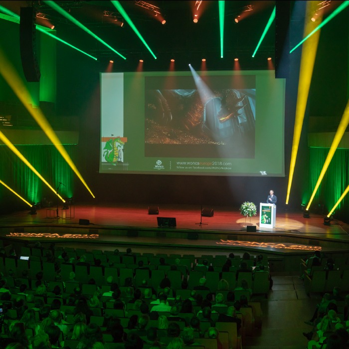Sala konferencyjna wypełniona publicznością podczas wydarzenia oświetlonego zielonymi i żółtymi reflektorami. Prelegent na scenie, duży ekran z prezentacją.