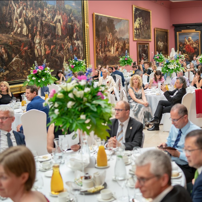 Elegancka kolacja galowa w muzeum, z gośćmi siedzącymi przy okrągłych stołach udekorowanych kwiatami. W tle obrazy w złotych ramach.