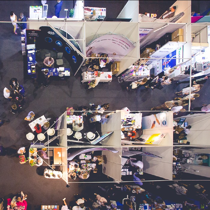 Widok z góry na stoiska targowe w Krakowie, z ludźmi spacerującymi między nimi. Różne ekspozycje i aranżacje stoisk, tworzące mozaikę kształtów i kolorów.