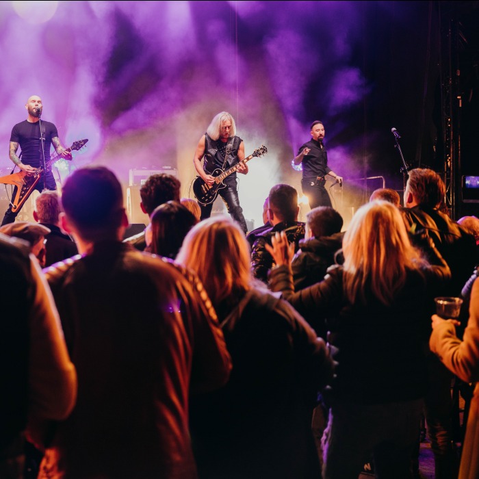 Zespół rockowy na scenie, oświetlony fioletowymi światłami, gra dla publiczności. Gitarzyści i wokalista w akcji, tłum widzów przed sceną.