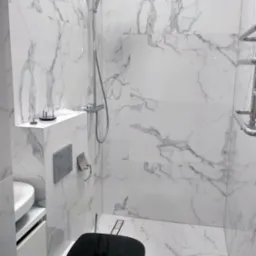 Nowoczesna łazienka z kabiną prysznicową typu walk-in, wykończona białym marmurem, z czarną deską sedesową i chromowaną armaturą. Minimalistyczny design.