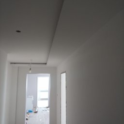 Lemberg Bud Sp. z o. o - Korytarz w trakcie wykończenia: gładkie, białe ściany, sufit z podwieszanym elementem i otwór drzwiowy bez ościeżnicy. W tle widoczny pokój z oknem.