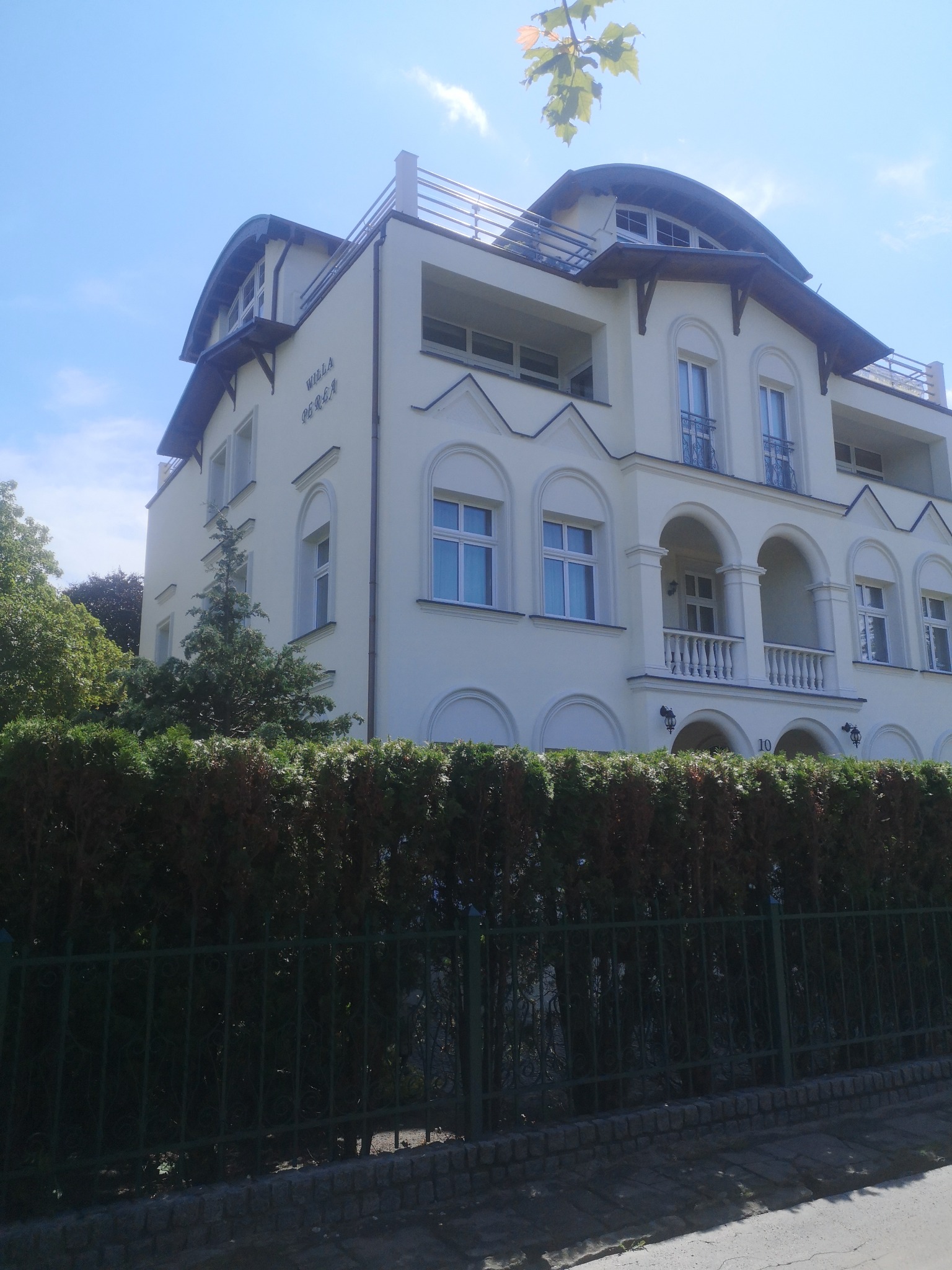Elewacja budynku Villa Perła z balkonami, widoczna zza żywopłotu i zielonego ogrodzenia. Jasna fasada z łukowymi oknami i balkonami w stylu architektonicznym.