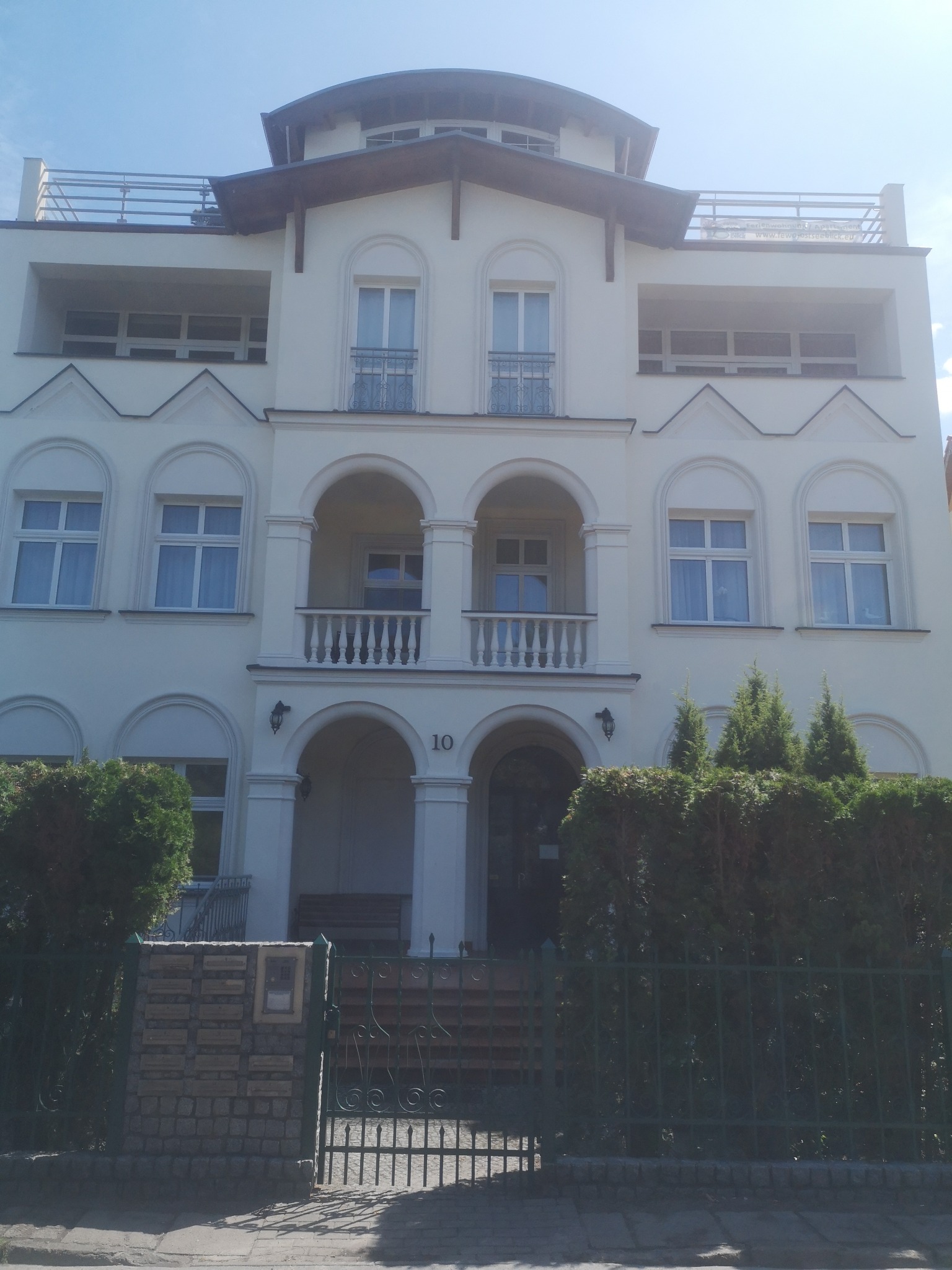 Elewacja trzypiętrowego budynku z balkonami i tarasem na dachu, utrzymana w jasnej kolorystyce. Widoczne liczne okna z dekoracyjnymi elementami architektonicznymi.