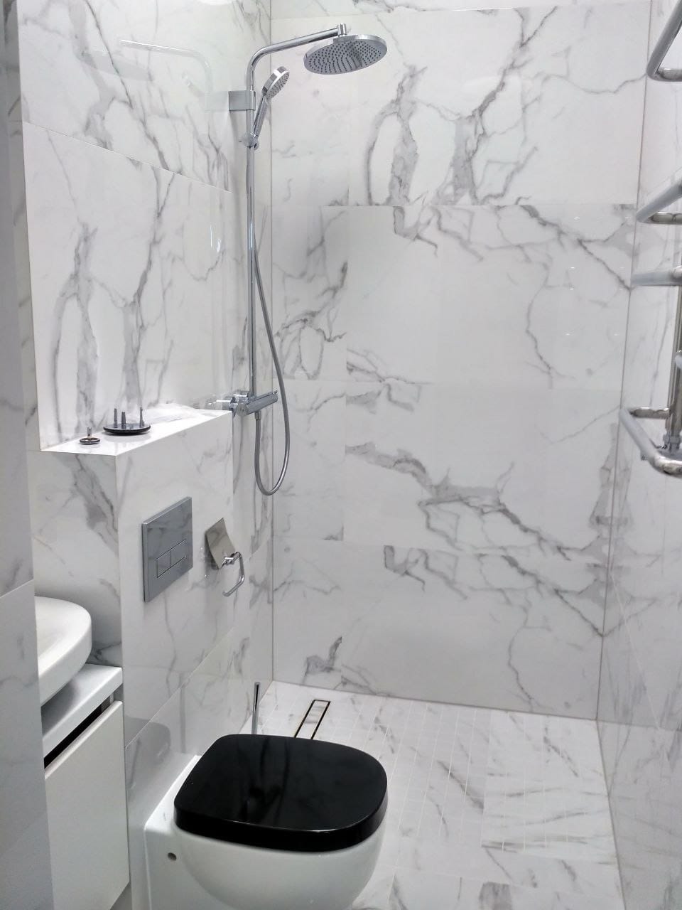 Nowoczesna łazienka z kabiną prysznicową typu walk-in, wykończona białym marmurem, z czarną deską sedesową i chromowaną armaturą. Minimalistyczny design.