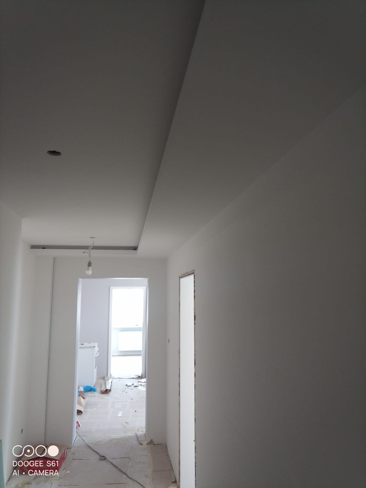 Korytarz w trakcie wykończenia: gładkie, białe ściany, sufit z podwieszanym elementem i otwór drzwiowy bez ościeżnicy. W tle widoczny pokój z oknem.