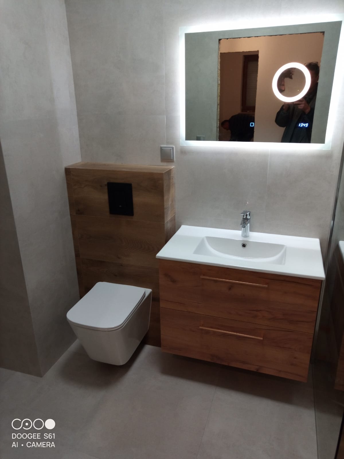 Nowoczesna łazienka z podwieszaną toaletą i umywalką w drewnianej szafce, podświetlane lustro z odbiciem osoby robiącej zdjęcie. Szare płytki na ścianach i podłodze.