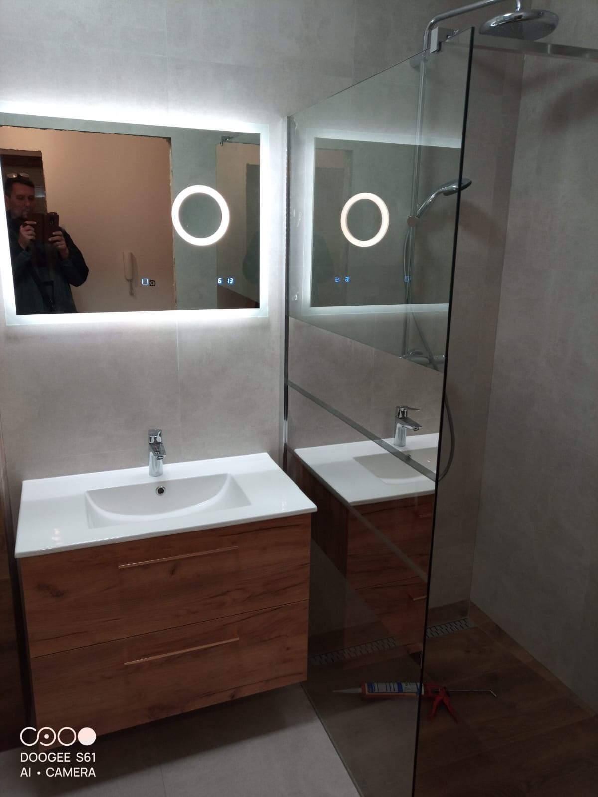 Nowoczesna łazienka po remoncie: umywalka z drewnianą szafką, podświetlane lustro i kabina prysznicowa ze szklaną ścianką. Widoczne narzędzia i silikon.