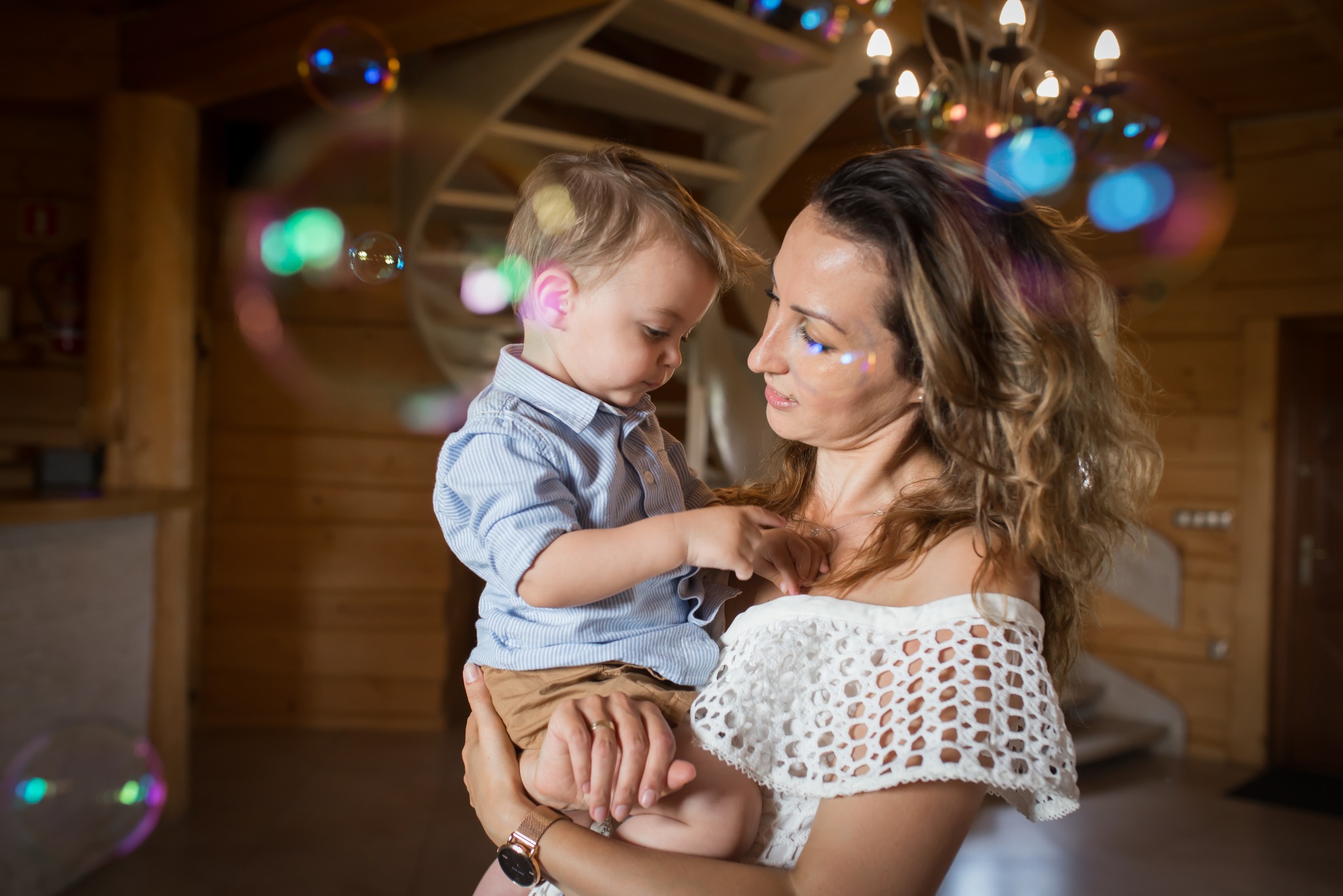 Matka z synkiem w objęciach, otoczeni bańkami mydlanymi w drewnianym wnętrzu. Kobieta ma brokat na twarzy, a w tle widoczny żyrandol i schody.