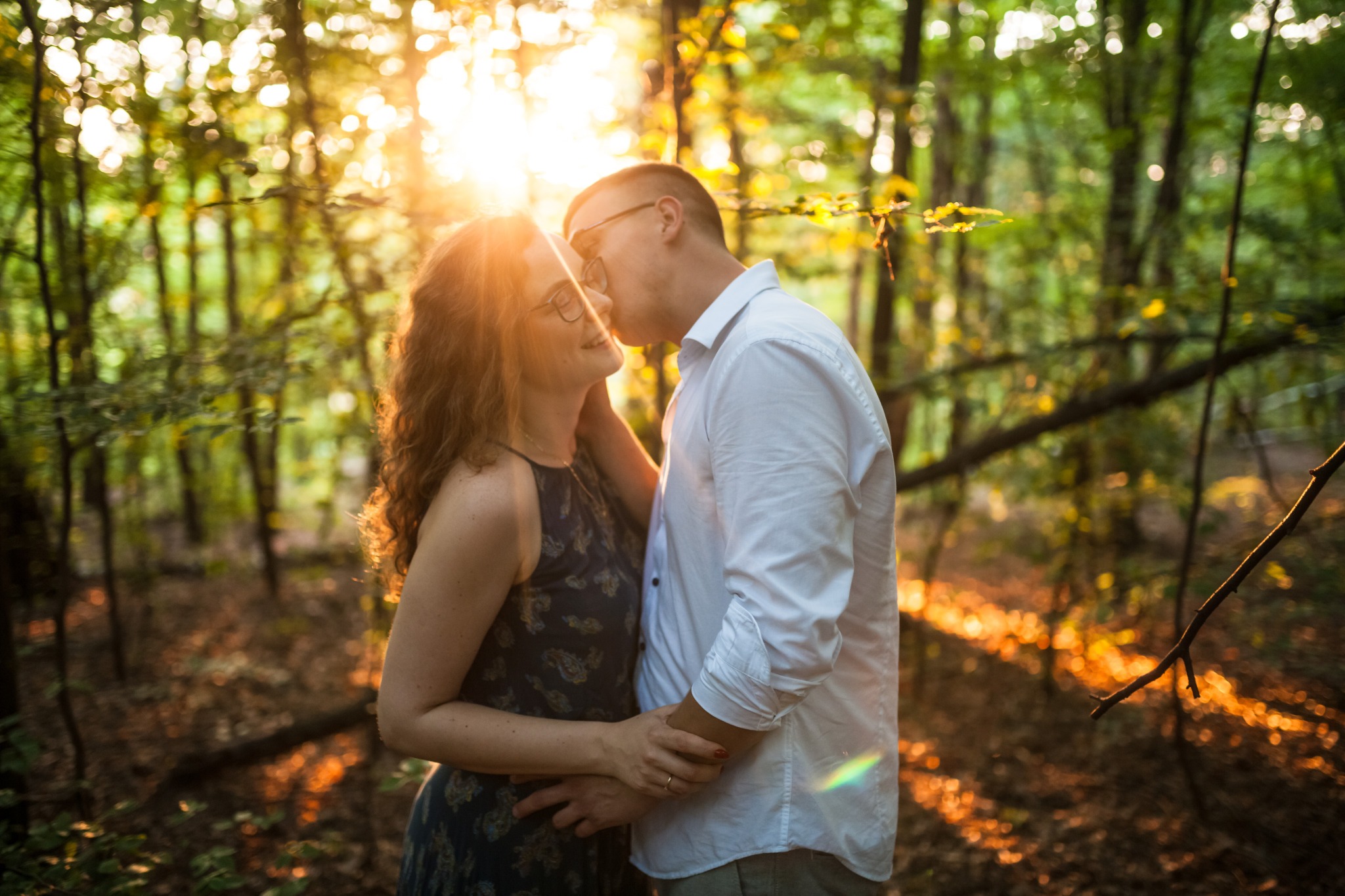 Zakochana para w okularach, w leśnej scenerii, delikatnie się całuje, otoczona promieniami zachodzącego słońca, tworząc romantyczny i intymny moment.