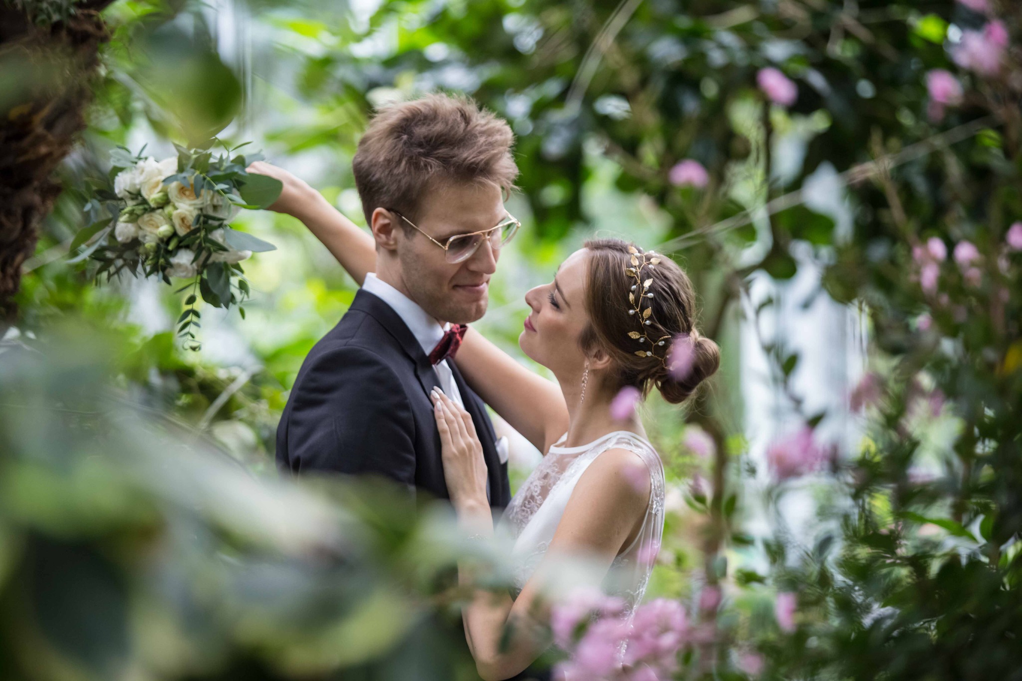 Para młoda w objęciach, on w garniturze z muszką, ona w sukni ślubnej z kwiatami we włosach, otoczeni zielenią i różowymi kwiatami w plenerze.