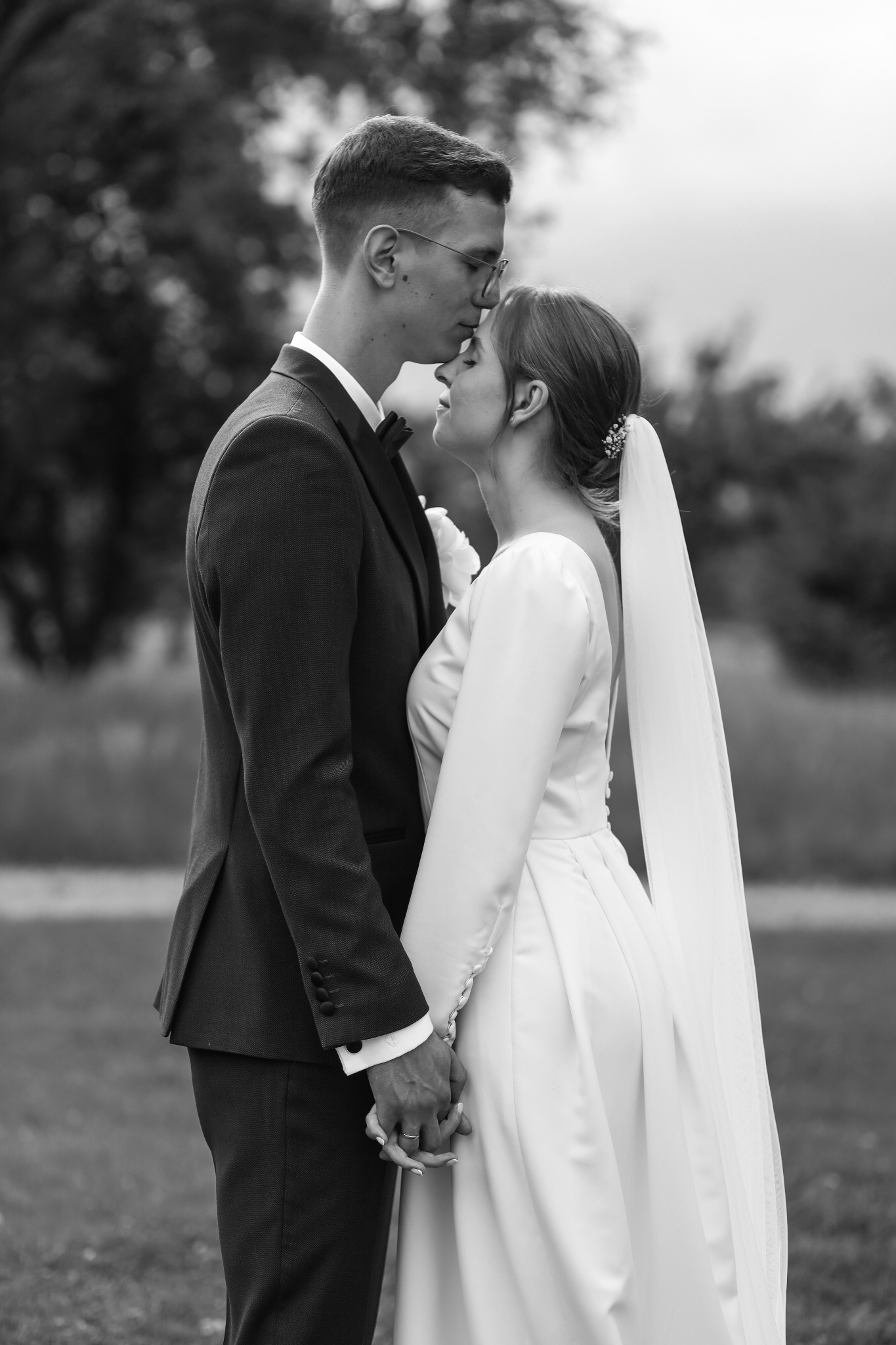 Czarno-białe zdjęcie pary młodej: pan młody w okularach całuje pannę młodą w czoło, trzymając się za ręce na tle zieleni.