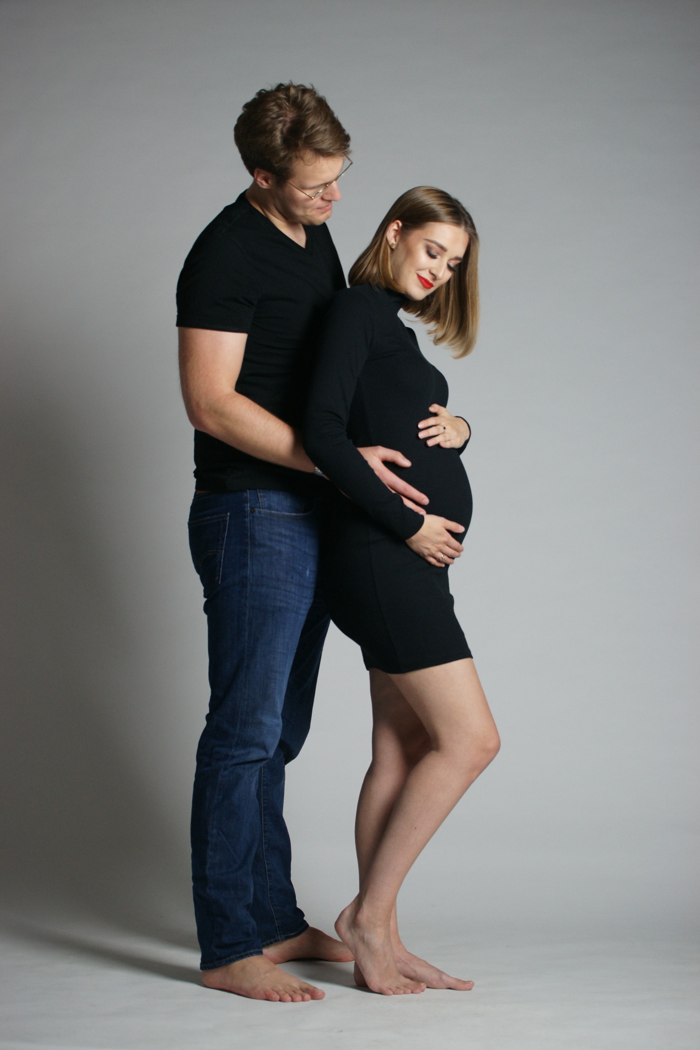 Sesja ciążowa w studio: przyszła mama w czarnej sukience, czułe objęcie partnera, stonowane tło, naturalne światło, podkreślony makijaż, bose stopy.
