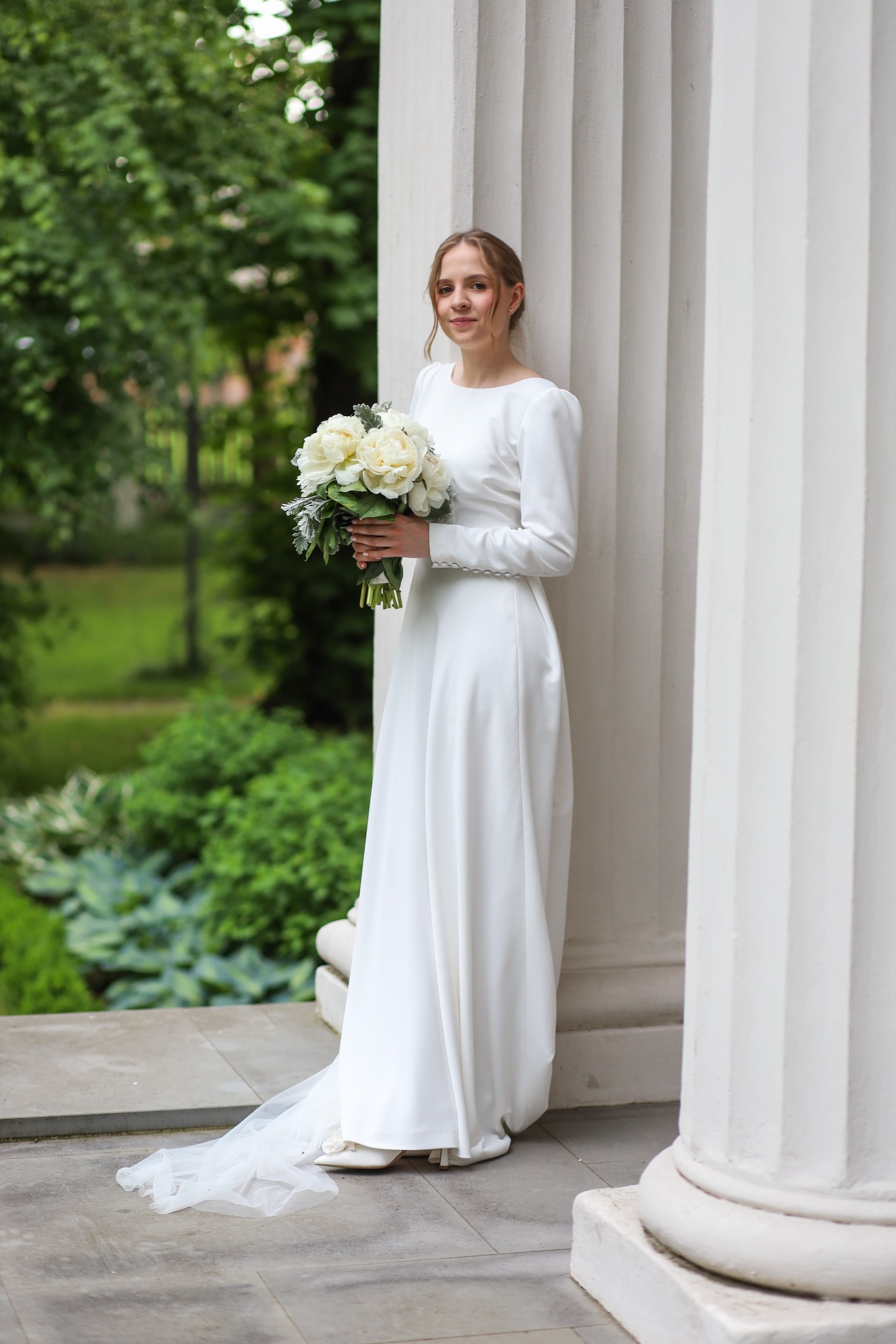 Panna młoda w prostej, białej sukni z długim rękawem, trzymająca bukiet białych kwiatów, pozuje przy kolumnie na tle zieleni.