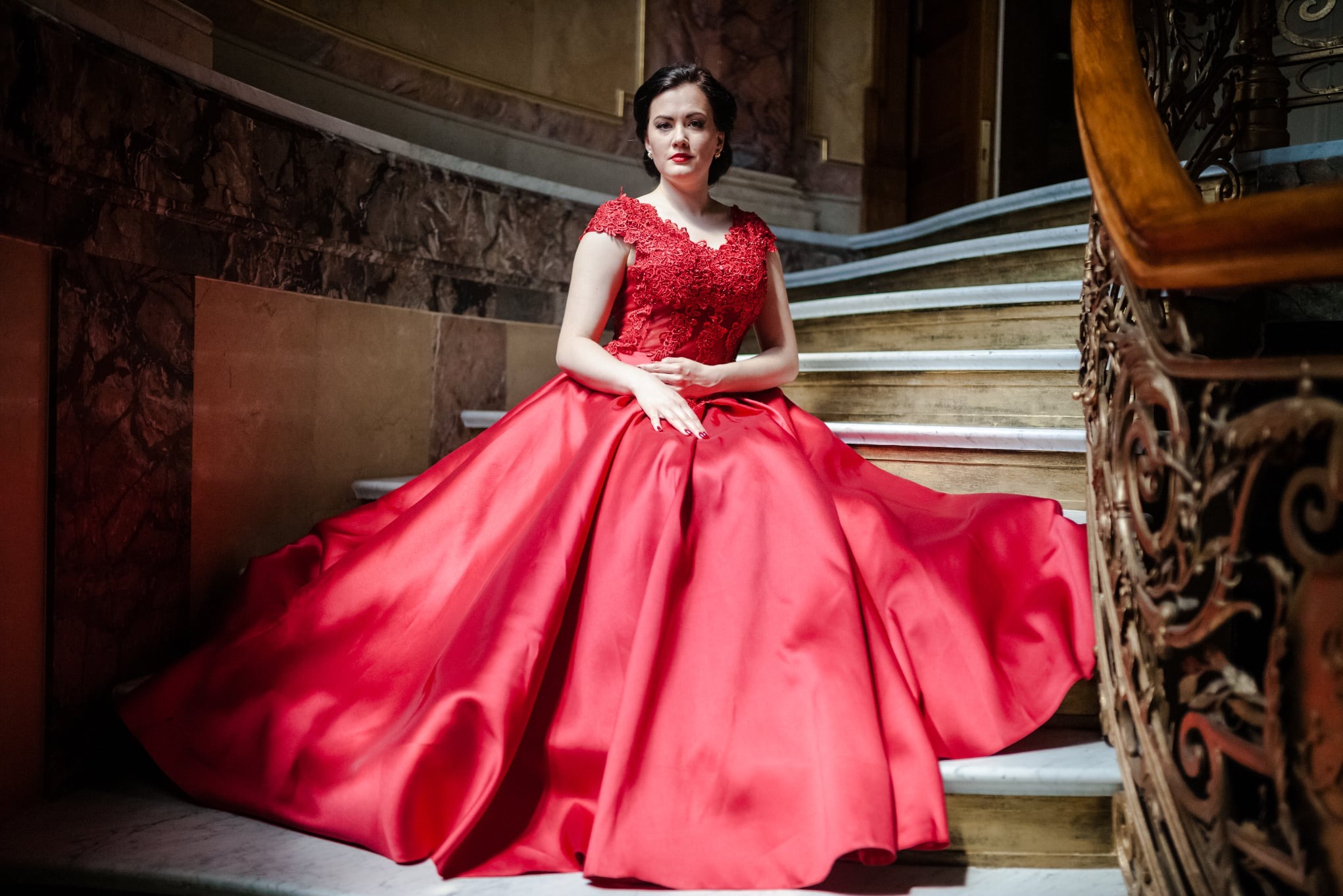 Kobieta w czerwonej, balowej sukni siedzi na marmurowych schodach z ozdobną, metalową balustradą. Ujęcie z dołu podkreśla elegancję kreacji i otoczenia.